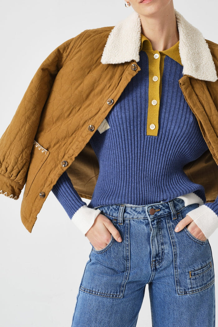 Person wearing a brown jacket with a white collar, blue ribbed sweater, and blue jeans