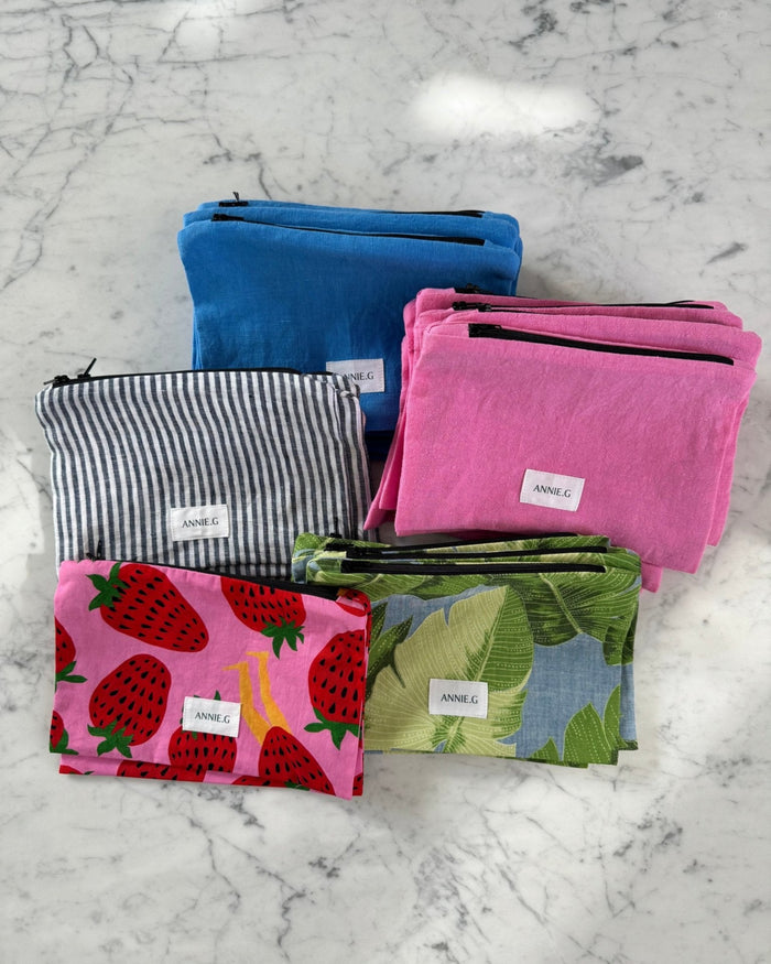 Five stacks of different coloured fabric zip top Pouches with ecru ANNIE.G brand tag sewn on the front - ANNIE.G