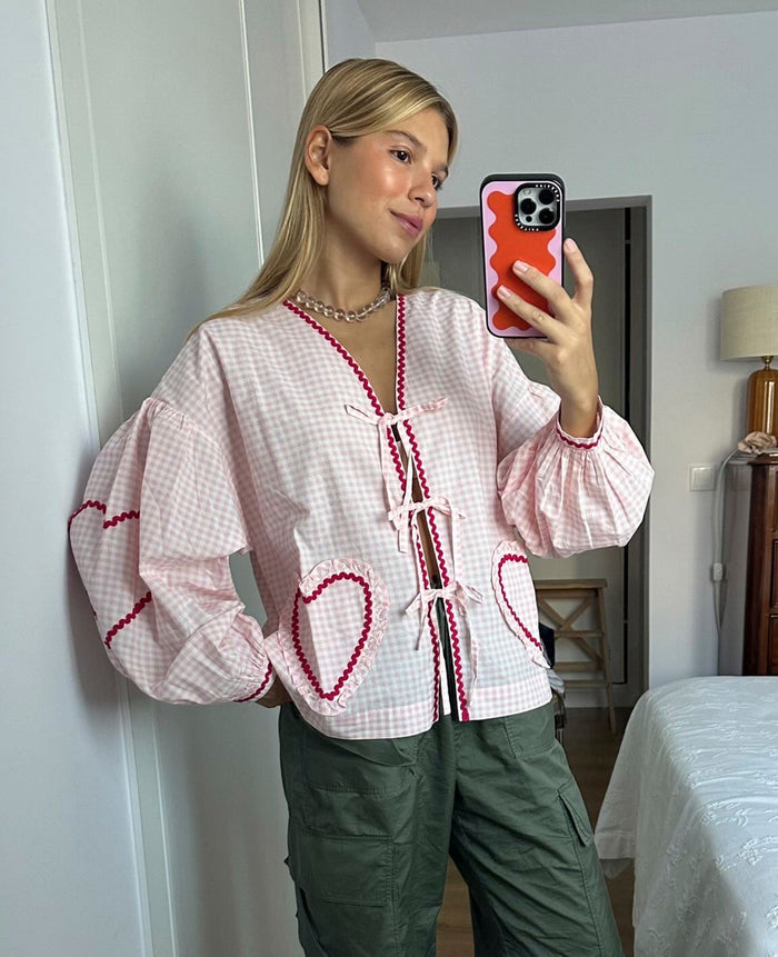 Woman taking a selfie and wearing long sleeve pink checked blouse with front tie closures and heart shaped pockets.