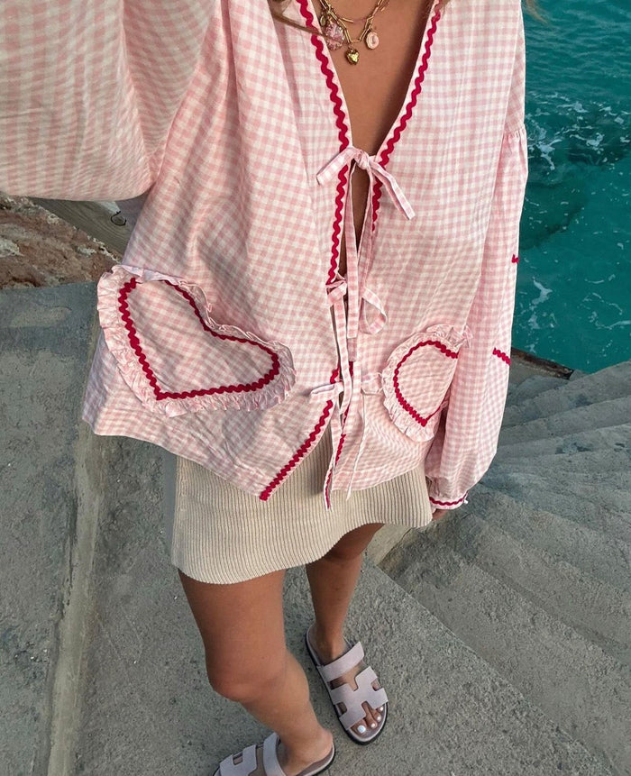 Close up of a woman wearing a long sleeve pink checked blouse with front tie closures and heart shaped pockets near water.