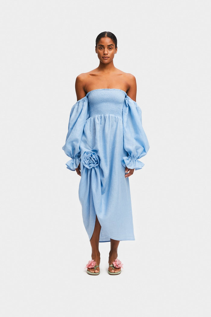 Woman wearing an off the shoulder blue midi dress with rose detail, smocked bodice and long sleeves with frill.