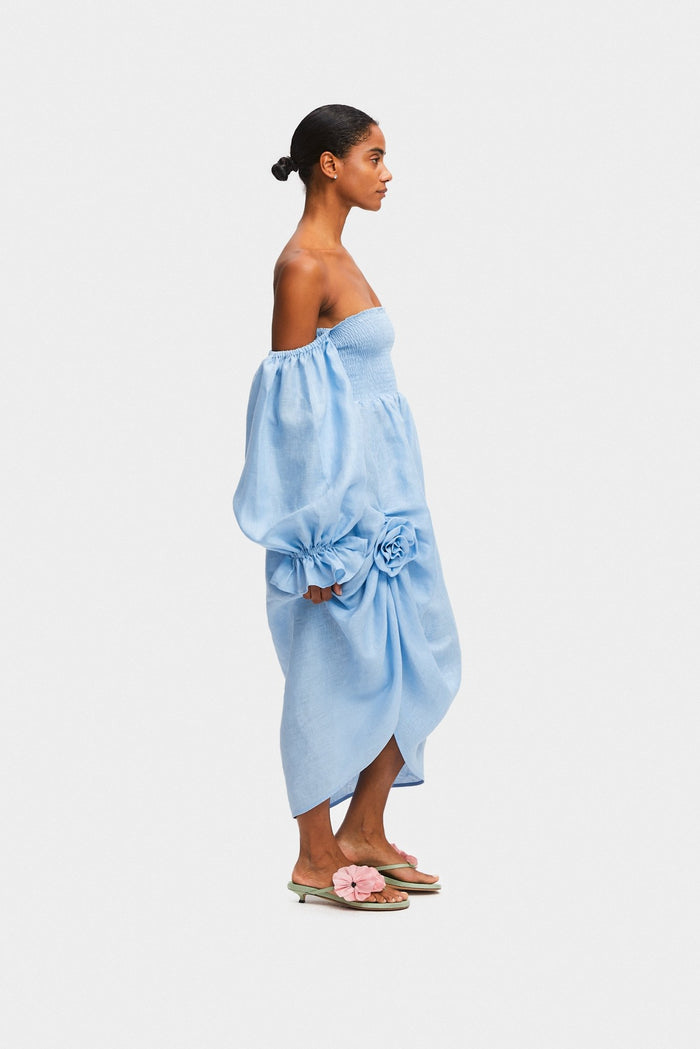 Woman standing on the side wearing an off the shoulder blue midi dress with rose detail creating a side split, smocked bodice and long sleeves with frill.