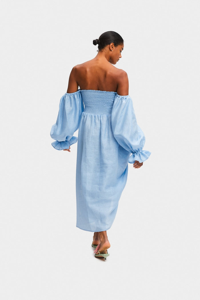 Back of a woman wearing an off the shoulder blue midi dress with a smocked bodice and long sleeves with frill.
