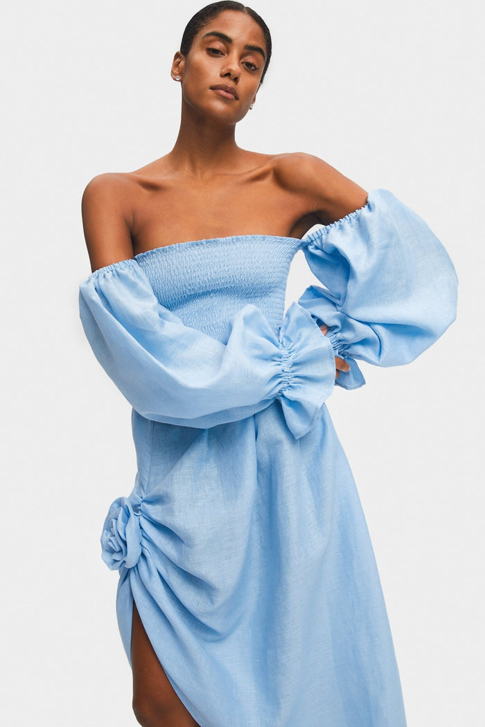 Woman wearing an off the shoulder blue midi dress with rose detail, smocked bodice and long puffed sleeves with frill.