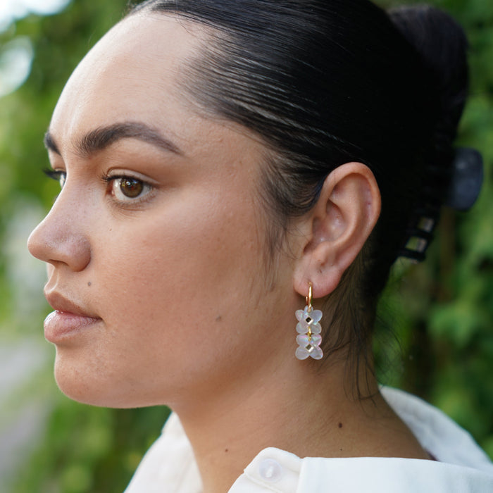Woman wearing dangle earring in shape of two sweet clover acrylic flowers inset with gold mirror diamond in white swirl acrylic on small gold hoops.