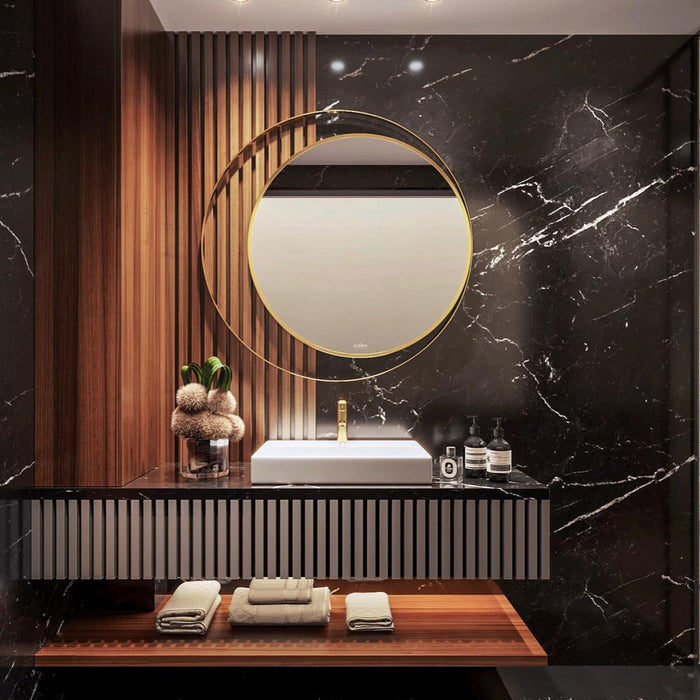 Modern and elegant bathroom vanity with a round mirror featuring a stainless steel gold colored frame and a sleek white marble sink countertop. With a plant and some perfumes kept on the counter top.