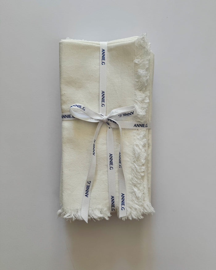 Folded and stacked frayed edge white cotton napkins with 'ANNE G' branding ribbon tied around.