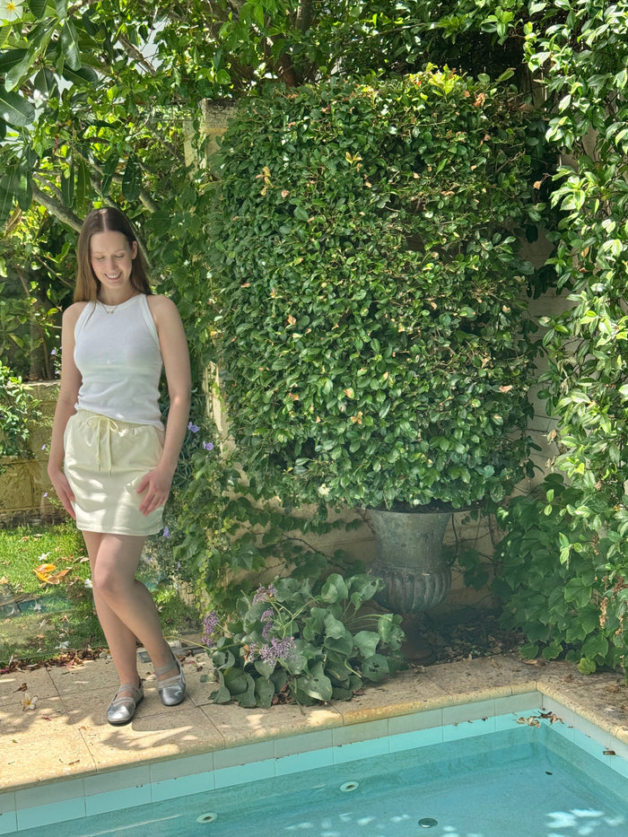 Woman standing by a pool with greenery wearing a light yellow miniskirt with pockets and tied drawcord with a white top and grey sandshoes.