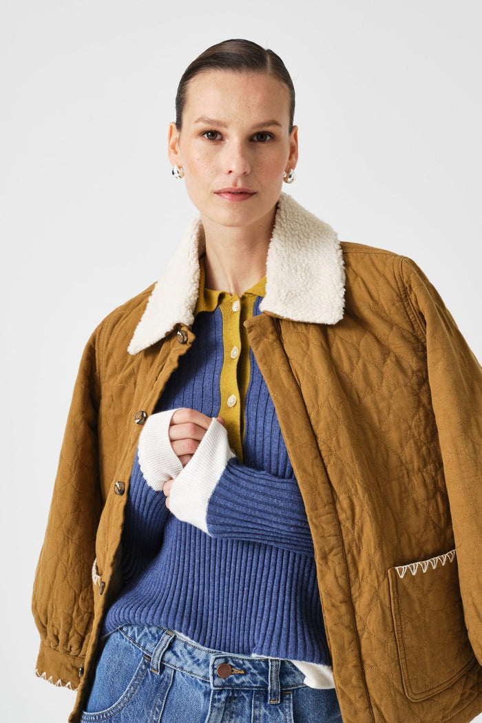 Woman wearing a brown quilted jacket with a white shearling collar, blue sweater, and jeans