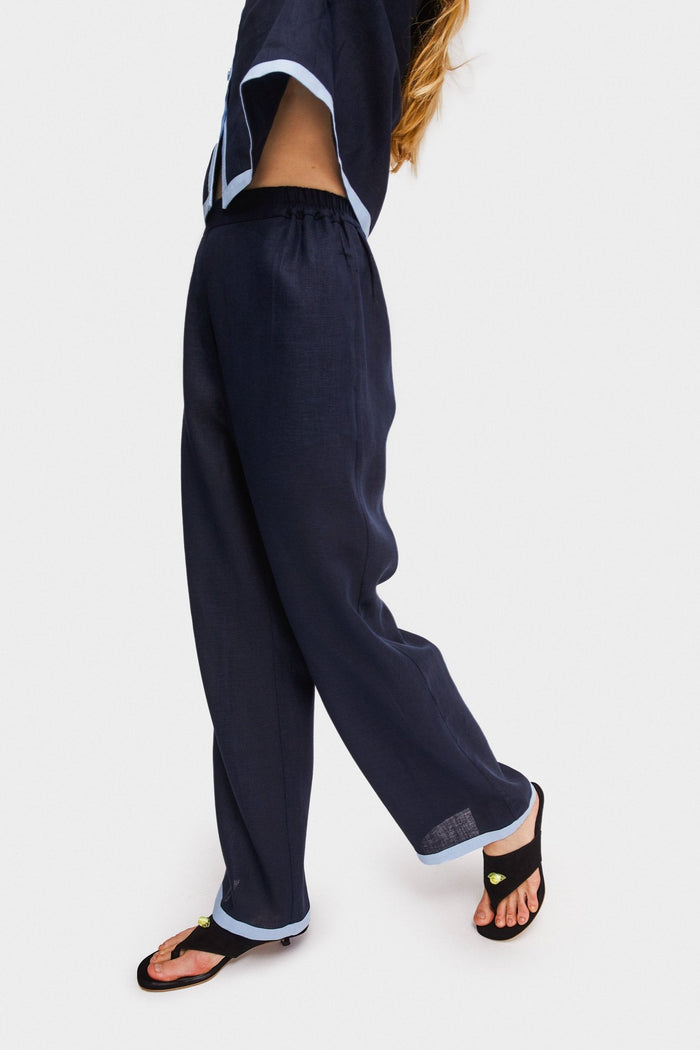 Woman standing on the side wearing long navy pants with contrast light blue trim on cuff and elastic waist band.
