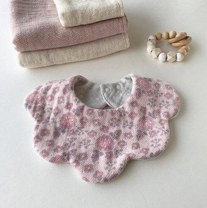 Pink floral-patterned baby bib with white dot jacquard reverse side in a scallop edge design and two snap closures lying next to towels and and wooden teething ring.