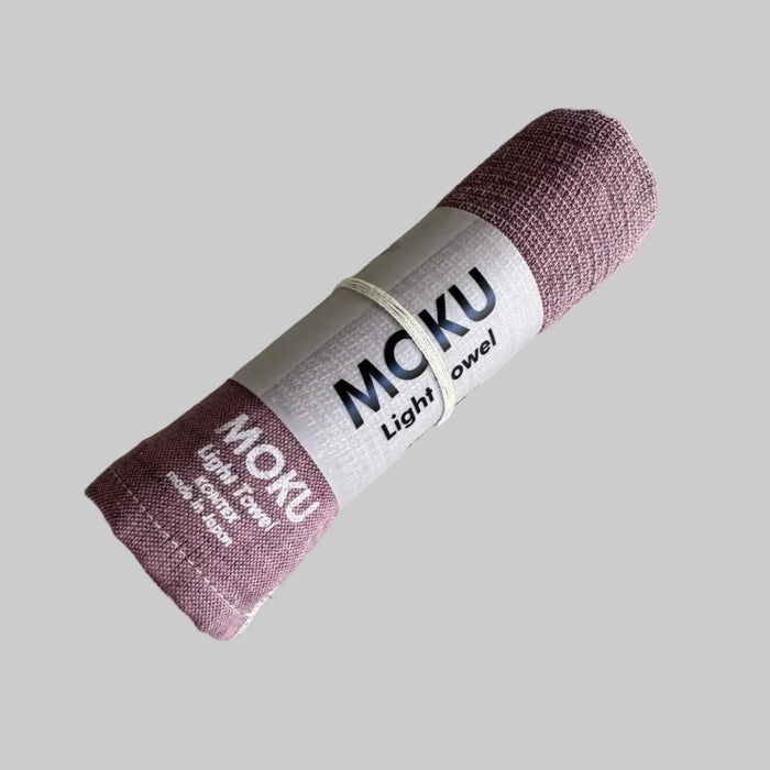 Pink coloured towel rolled in packaging sleeve with "MOKU Light Towel' written in white on fabric.