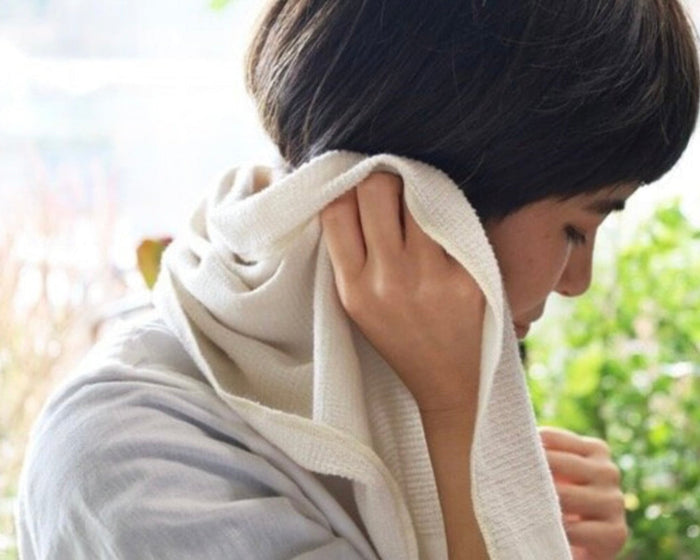 Person with short hair holding a towel to their face outdoors with greenery in the background.