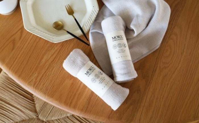 Two towels rolled with MOKU Organics label on a wooden table surface with a blueberry coloured towel, plate and cutlery.