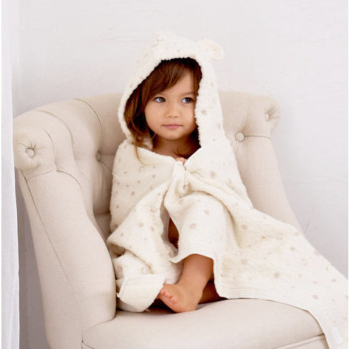 Child wrapped in a cream hooded towel sitting on a beige chair.
