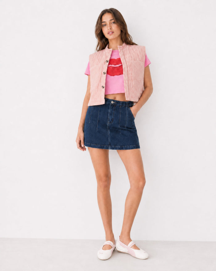 Woman wearing a pink quilted vest, pink crop top with red lips design on front, and denim mini skirt.