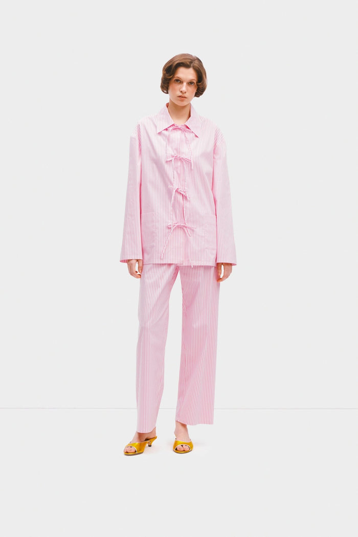 Woman wearing a Cotton-Poplin pink stripe pyjama set with long sleeve top and bow closure and pants.