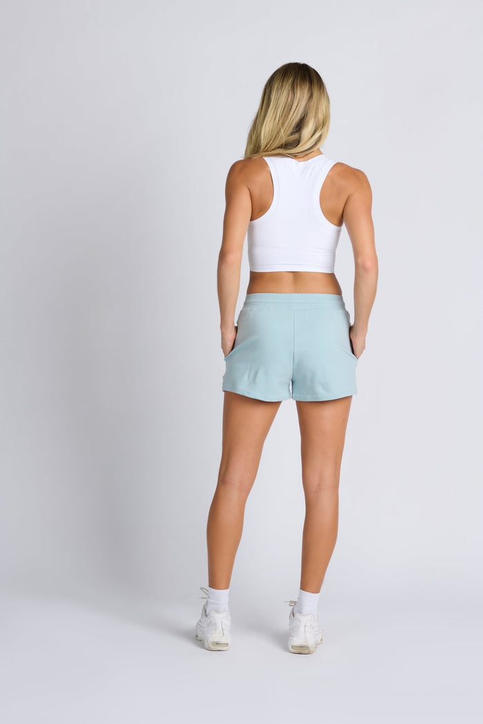 Back of a woman wearing light blue french terry sweat shorts with hands in pockets and white crop top with sporty racer back - ANNIE.G