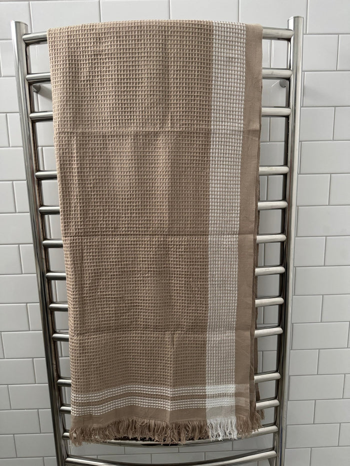 Bone with white border edge beach towel hanging over a towel rail in tiled bathroom.