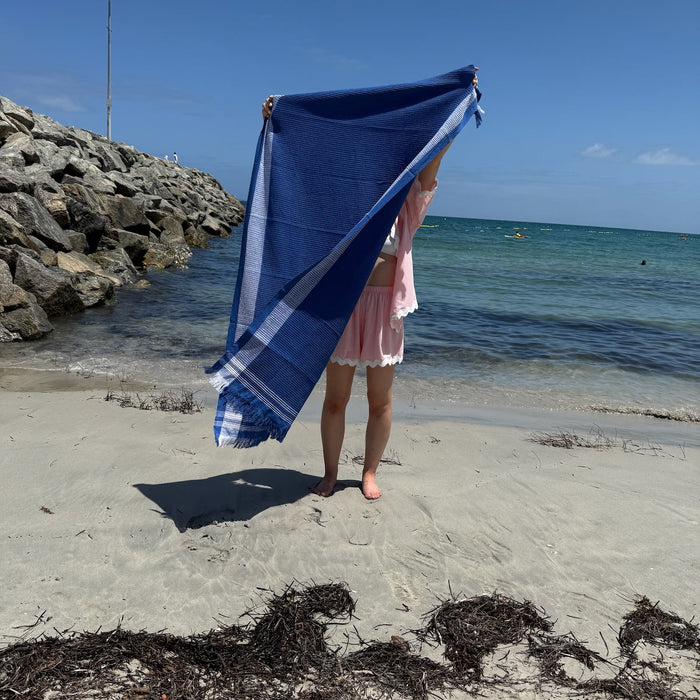 Woman holding up a navy waffle weave towel with white border and fringe ends at the beach - ANNIE.G