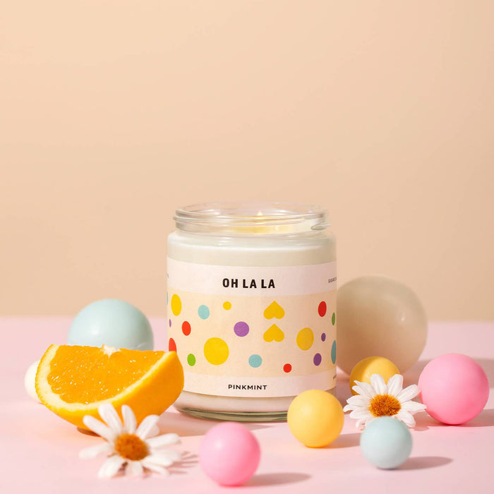 White soy candle in a jar with a wick lit and an orange sliced and flowers around it on a table - ANNIE.G