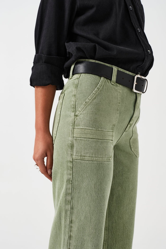Close up of the side of a person wearing green jeans with retro patch pockets and a black belt through loops with a black shirt.