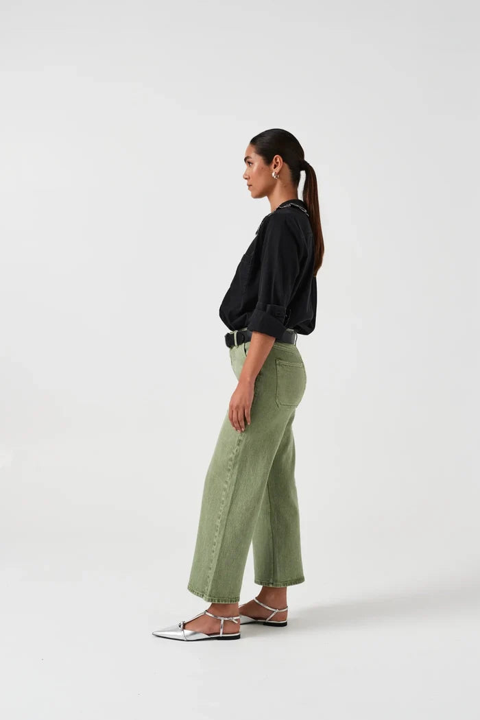 Woman wearing green cropped  jeans, a black shirt and sandals standing on the side.