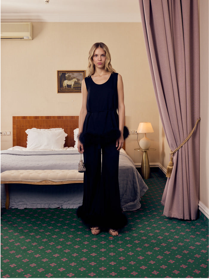 Woman in a black long pants and sleeveless top with feather trim and matching waist belt outfit standing in a bedroom.