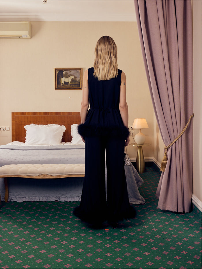 Woman in a black top with belt and long pants with black feather trim standing in a hotel room with a bed and lamp.