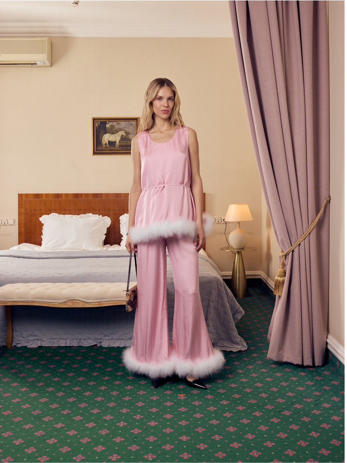 Woman in a pink sleeveless top with matching belt and pants with white feather trim standing in a bedroom.