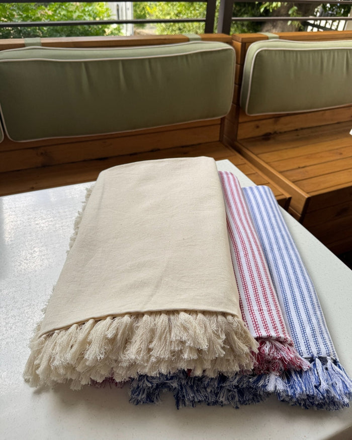 Stack of tablecloths folded on a table in natural, red ticking and blue ticking with fringe edge.