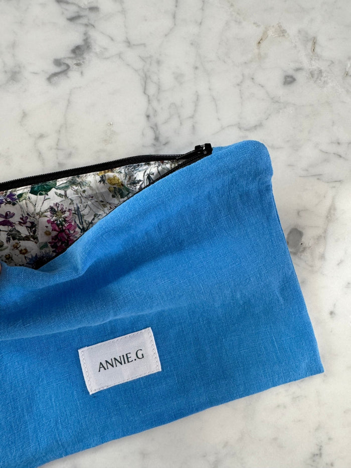 Cornflower blue linen pouch with top zip open showing floral lining inside and an ecru ANNIE.G logo tag sewn on the front lying flat on a bench - ANNIE.G