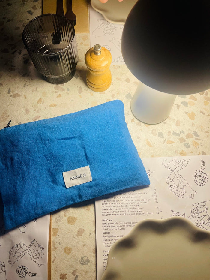Cornflower blue linen pouch with top zip closure and an ecru ANNIE.G logo tag sewn on the front lying flat on a cafe table under a table lamp - ANNIE.G